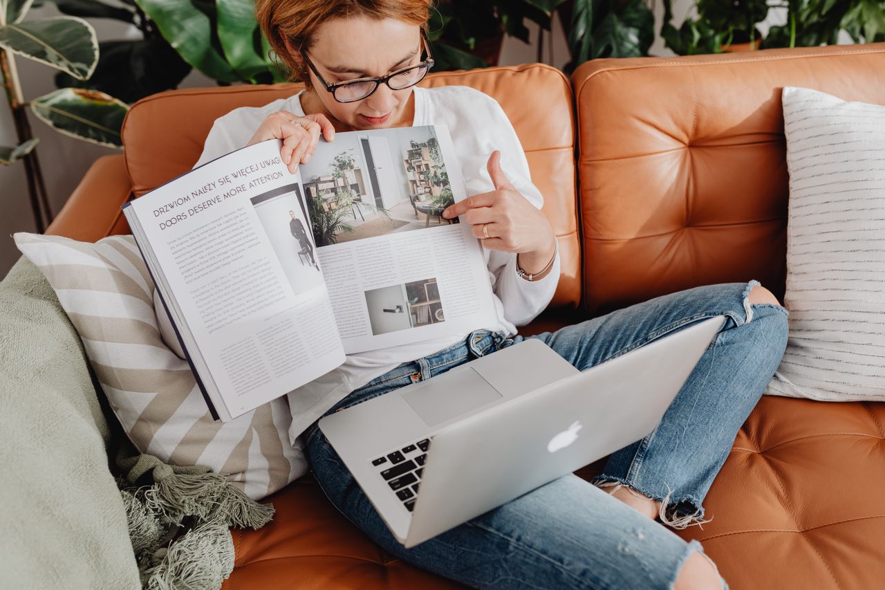 Femme lisant un magazine sur un canapé en cuir marron tout en utilisant un ordinateur portable.